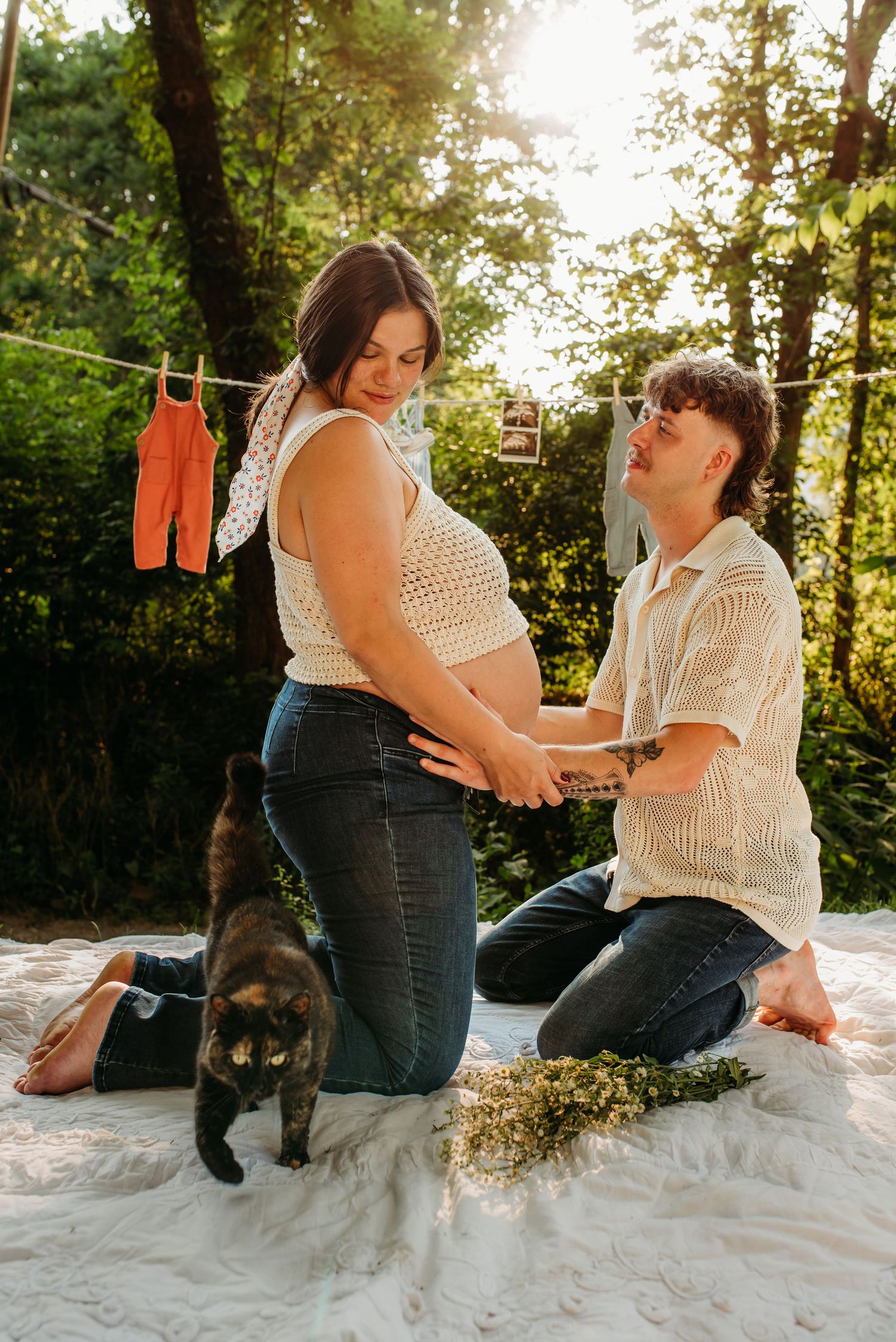 A couple sits on a white blanket in a forest setting during a romantic outdoor photoshoot.