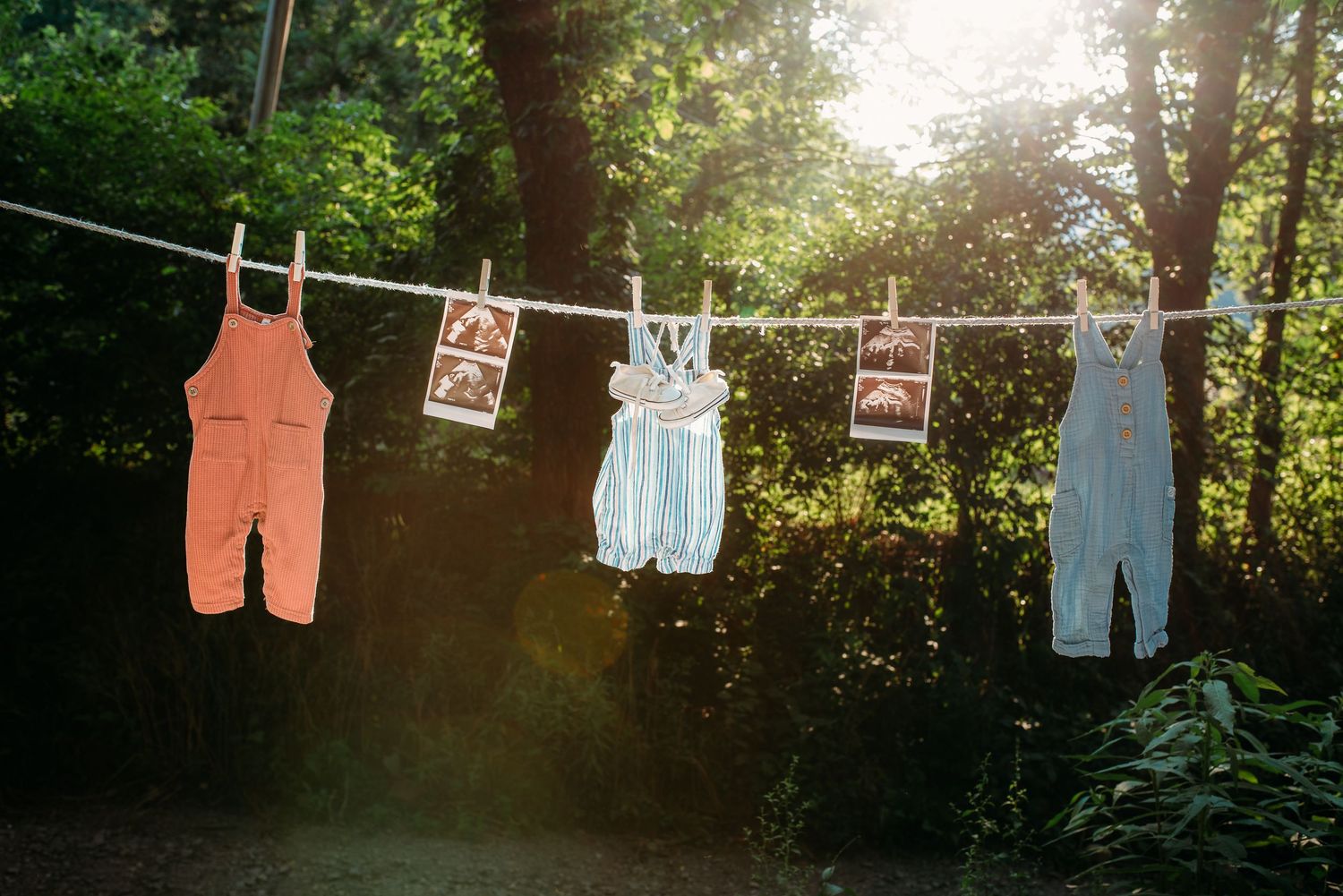 Baby clothes and overalls hanging on a clothesline at sunset creating a nostalgic silhouette against trees.