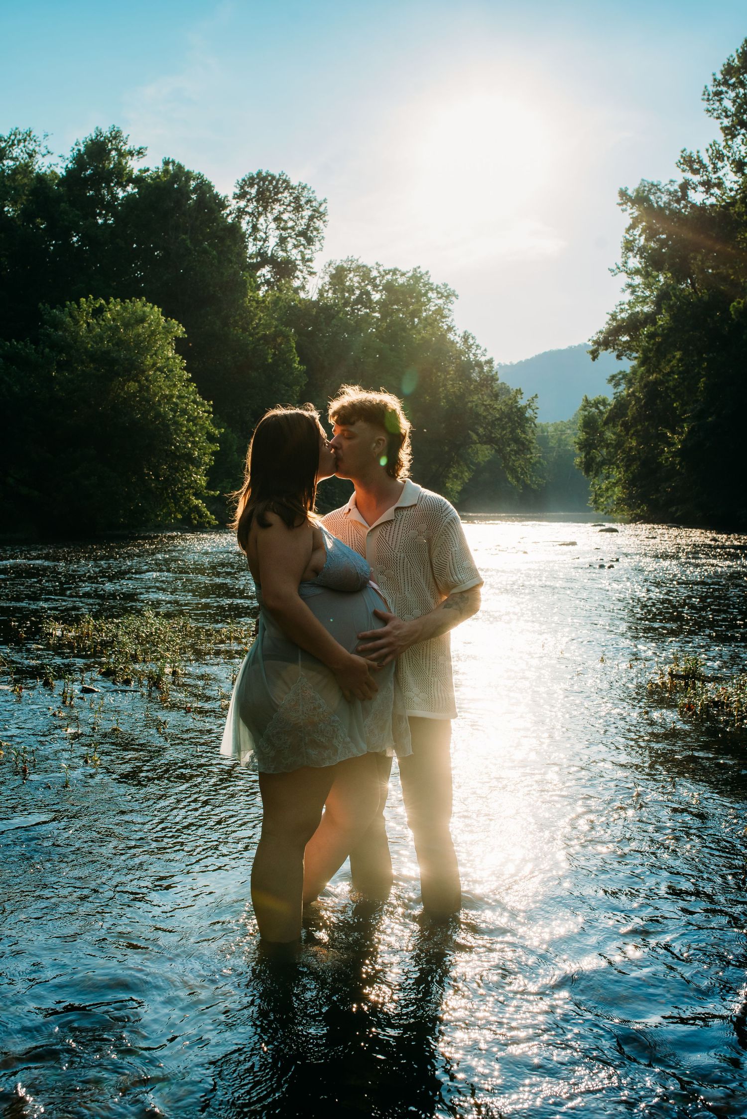 Silhouetted maternity photo of couple standing in flowing water at sunset with mountains in background.