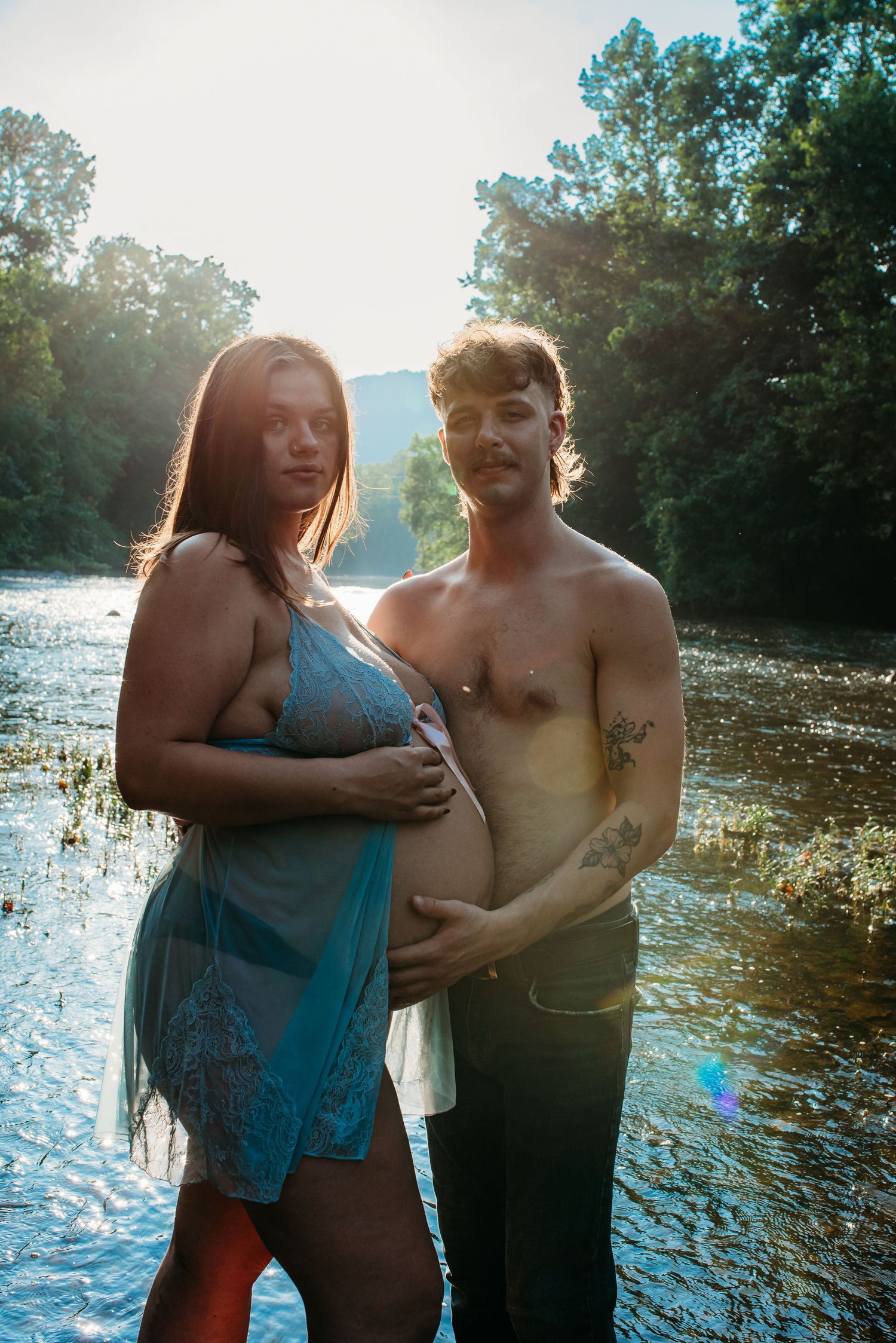 A maternity photo session in a river at golden hour with sun rays filtering through trees.