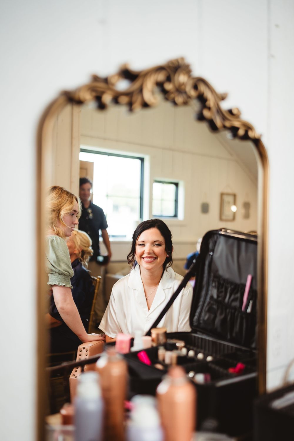 Mirror reflection shows beauty preparations with makeup case and ornate vintage frame in getting ready room.