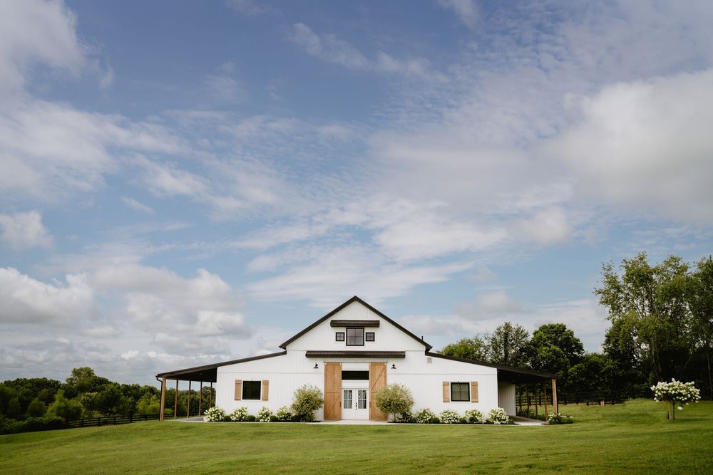 Modern white farmhouse wedding venue with large front door and green lawn under partly cloudy sky.