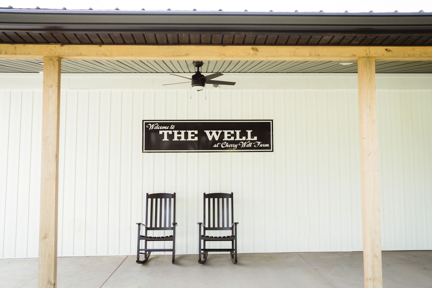Two wooden rocking chairs sit beneath a sign reading 'The Well' on a white porch with a ceiling fan.