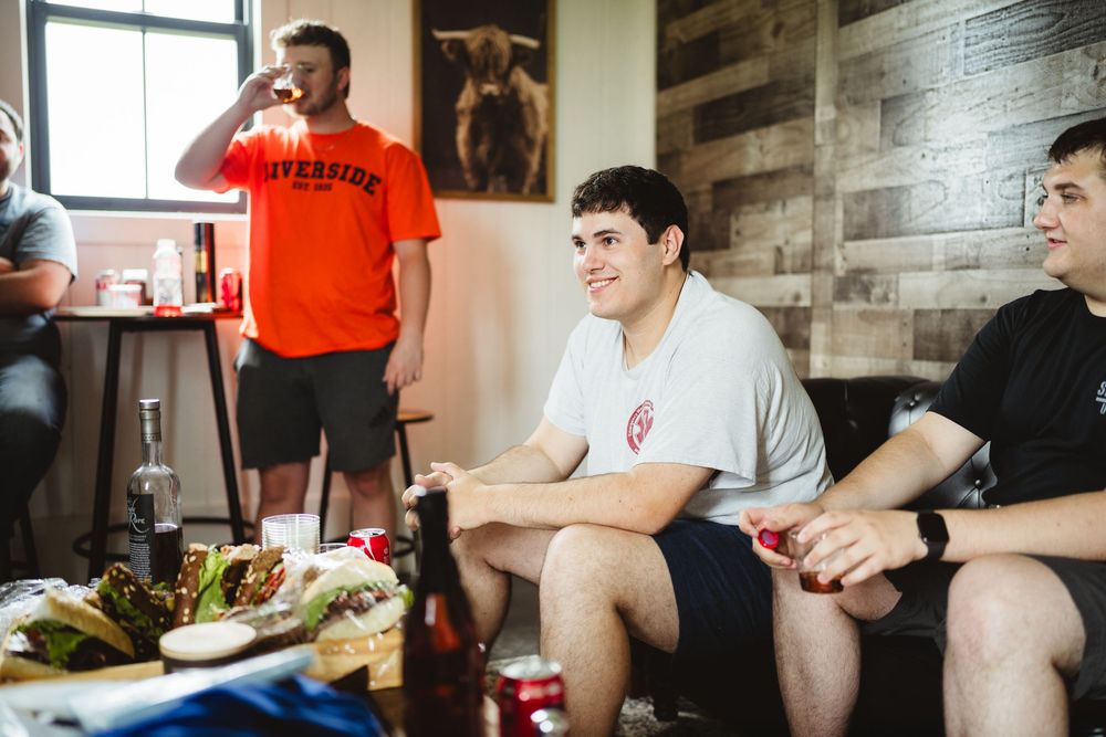 People relaxing on a couch with snacks and drinks in a room with rustic wood panel walls.
