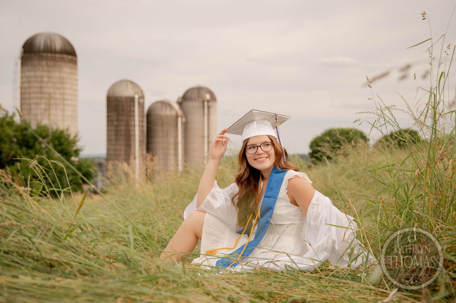 A figure in a white dress sits in tall grass near farm silos on a cloudy summer day.