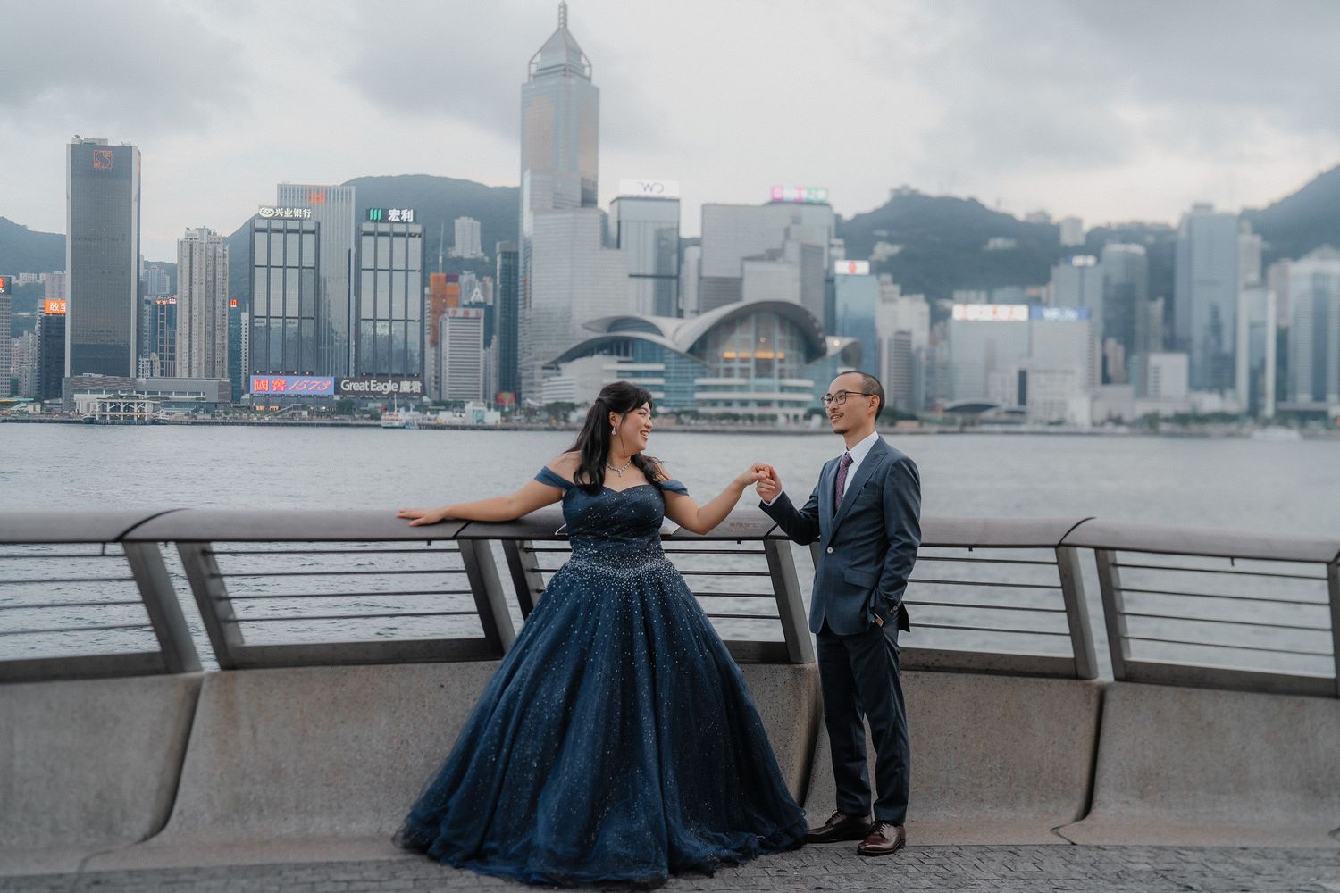 Two people in formal attire pose along Victoria Harbour with Hong Kong's iconic skyline and convention center in the background.