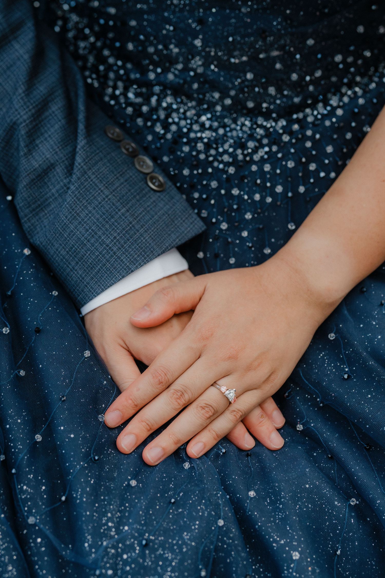 Close-up of hands clasped together showing an engagement ring against a sparkly navy blue dress.