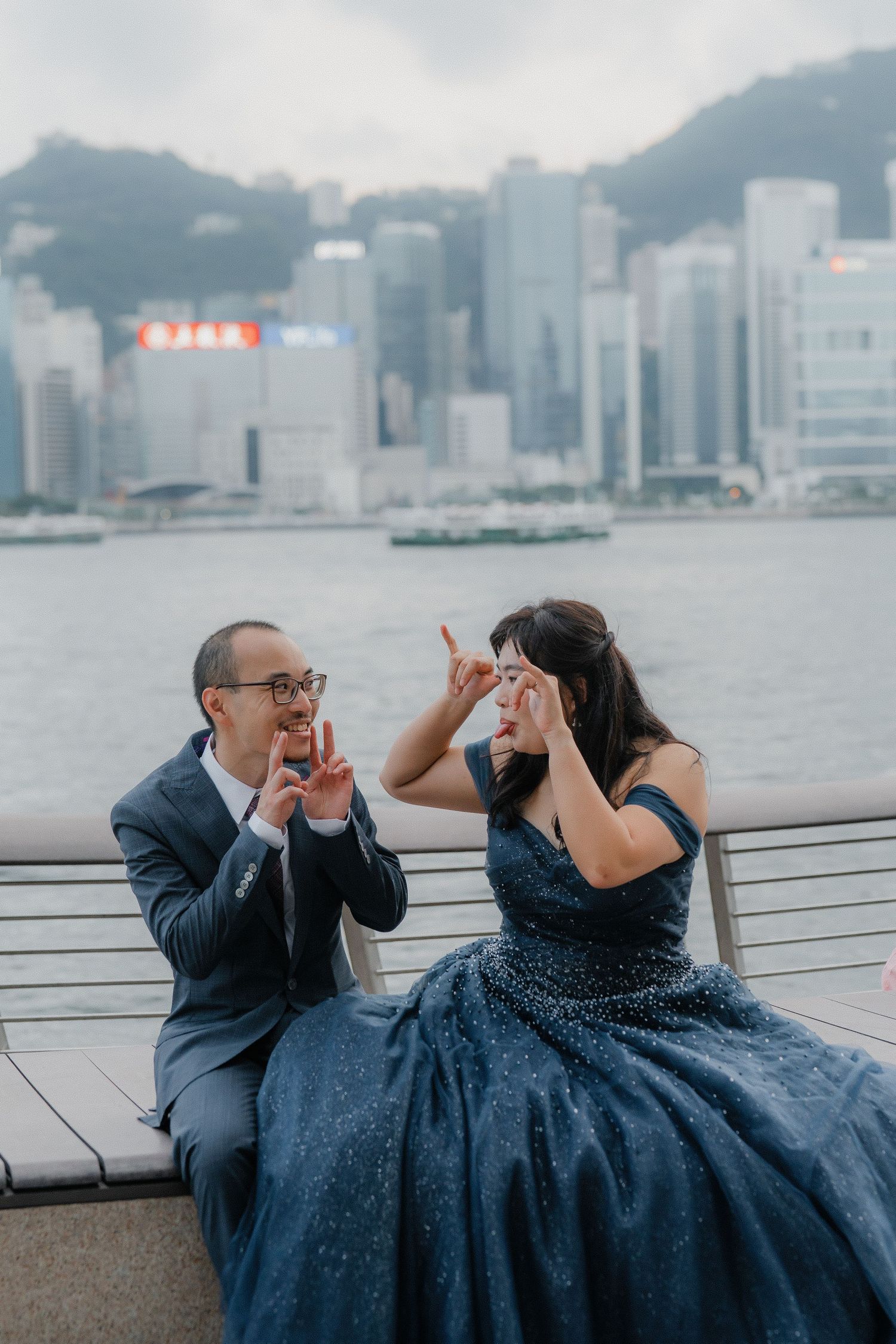 A playful couple posing by the waterfront with Hong Kong's harbor and skyline visible behind them.
