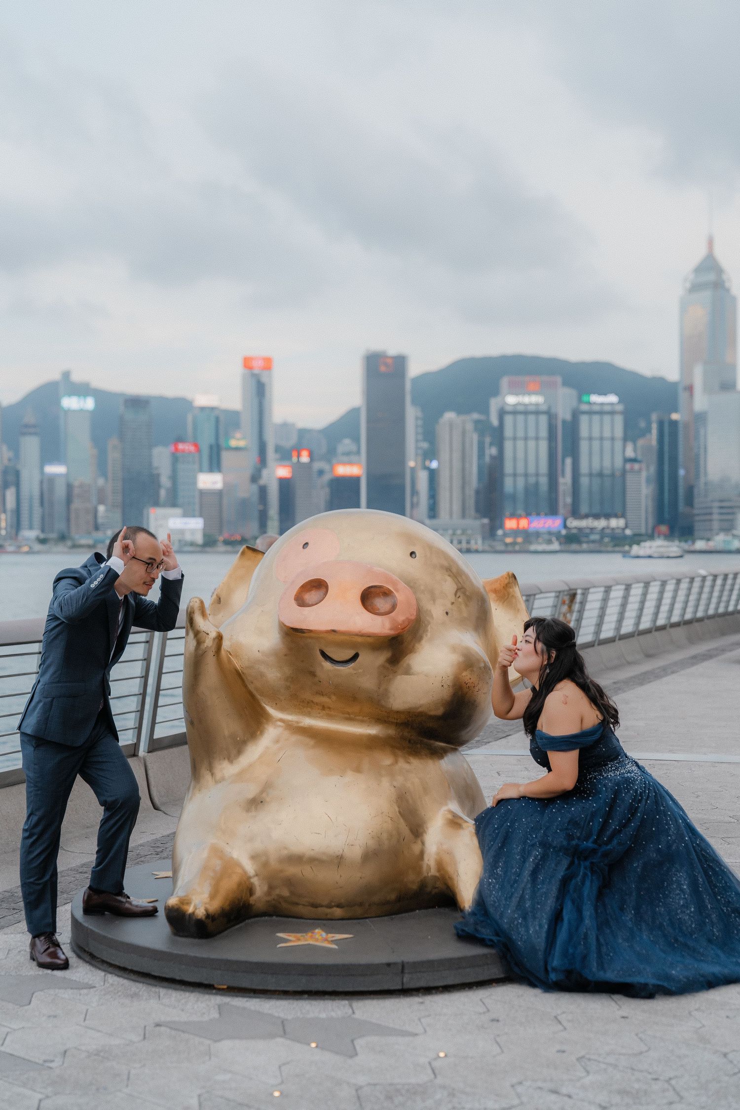 A couple poses playfully with a golden pig statue along Hong Kong's Victoria Harbour waterfront.