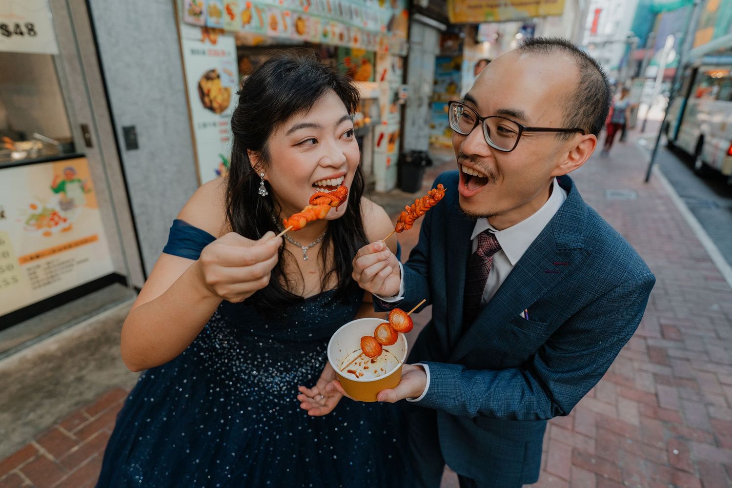 A couple enjoys street food together against a backdrop of colorful Chinese restaurant signs.