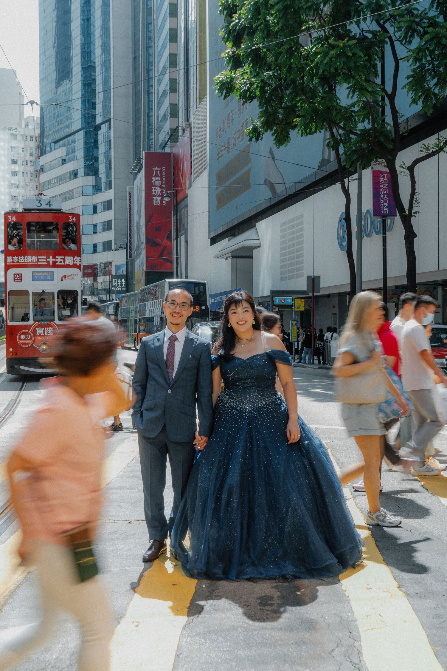 Couple in formal attire crossing a busy Hong Kong street with a red tram in the background.