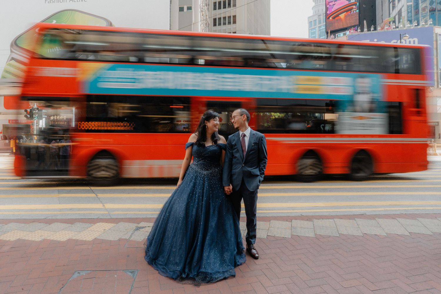 Motion blur photo of couple on Hong Kong street with red double-decker buses passing by.