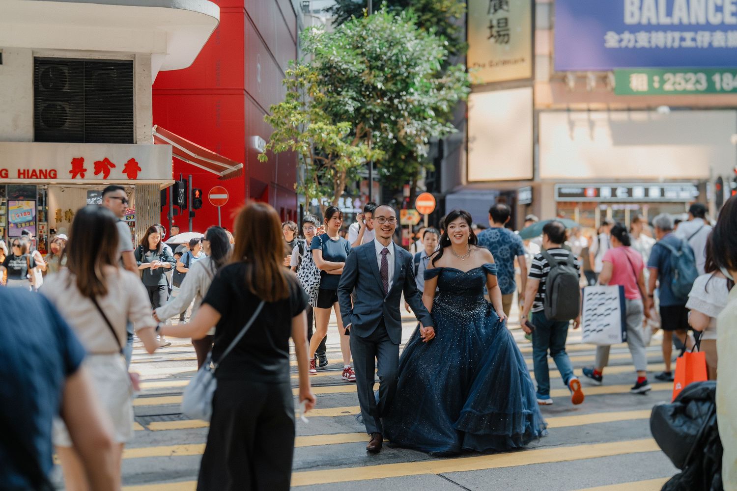 A couple poses playfully in front of a busy urban shopping district with storefront signs.