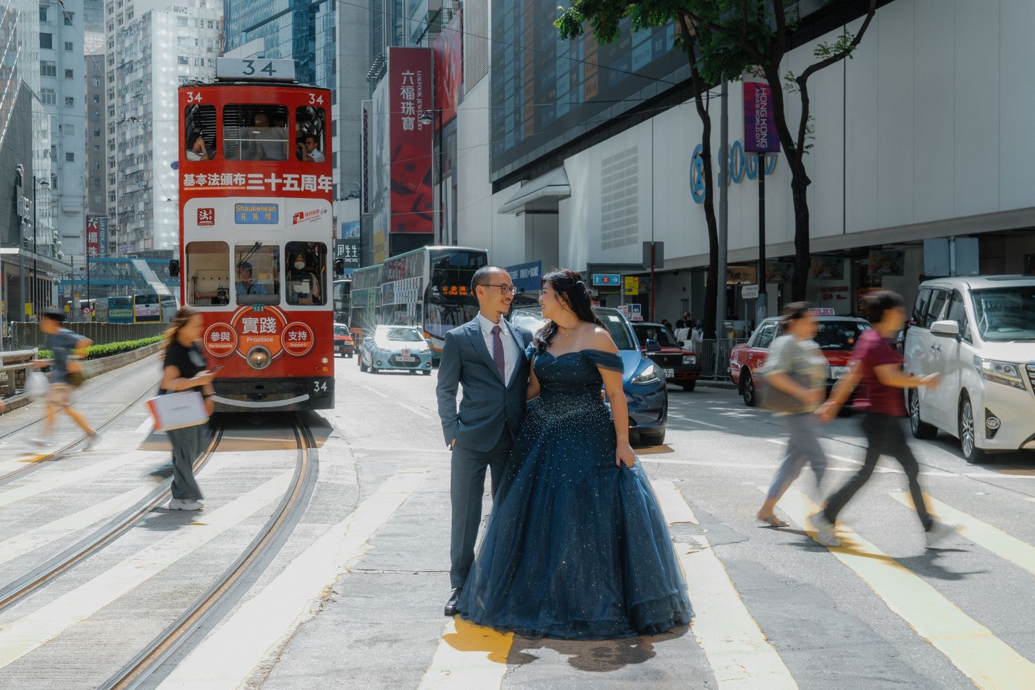 A couple walks through the busy streets of Hong Kong with classic trams and buildings in view.