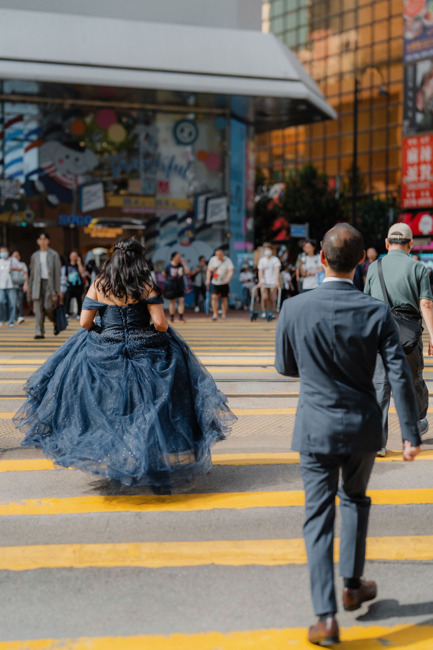 A person in a flowing blue gown crosses a busy street intersection while pedestrians wait at a crosswalk.