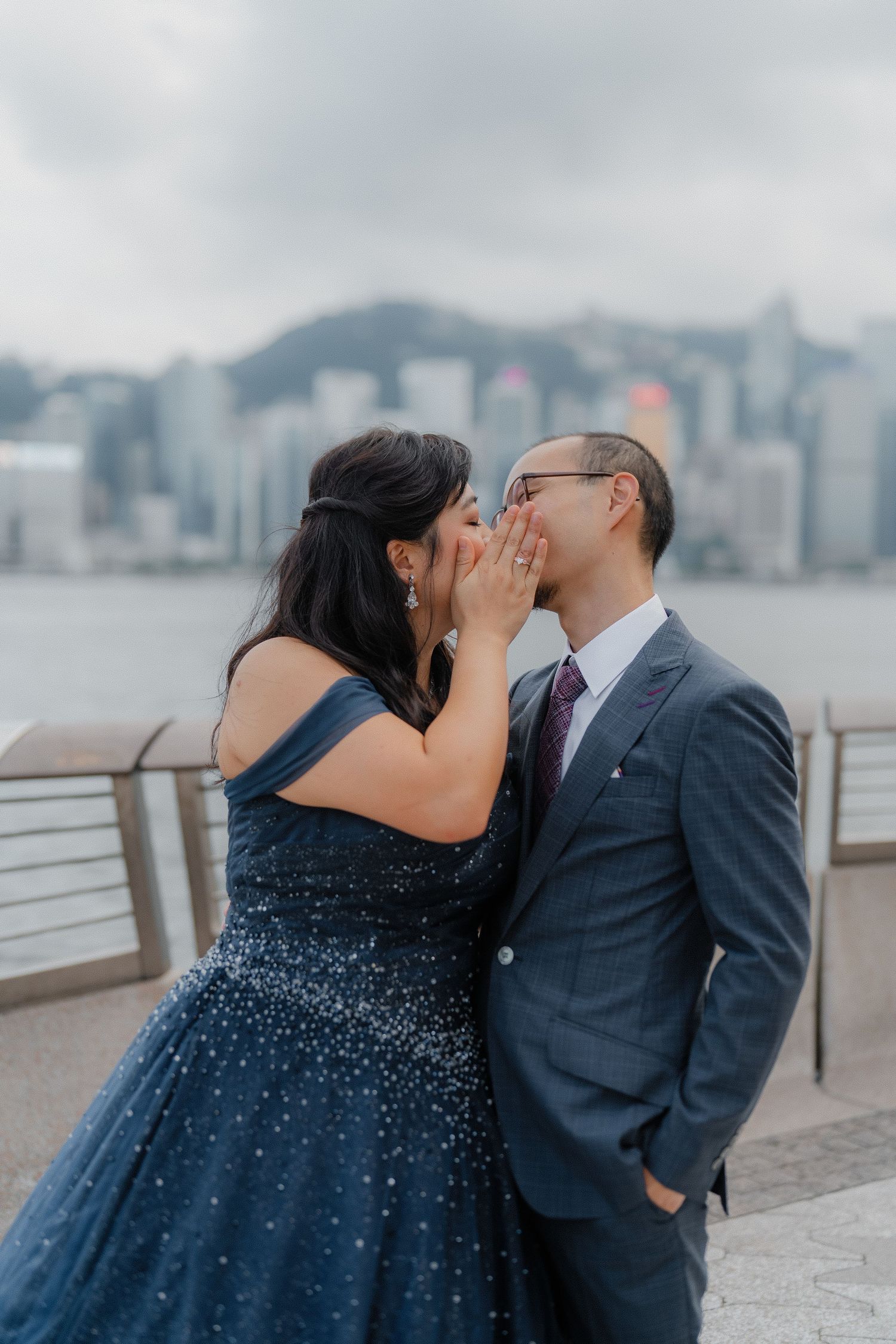 A couple shares an intimate moment on a waterfront promenade with Hong Kong's skyline in the misty background.