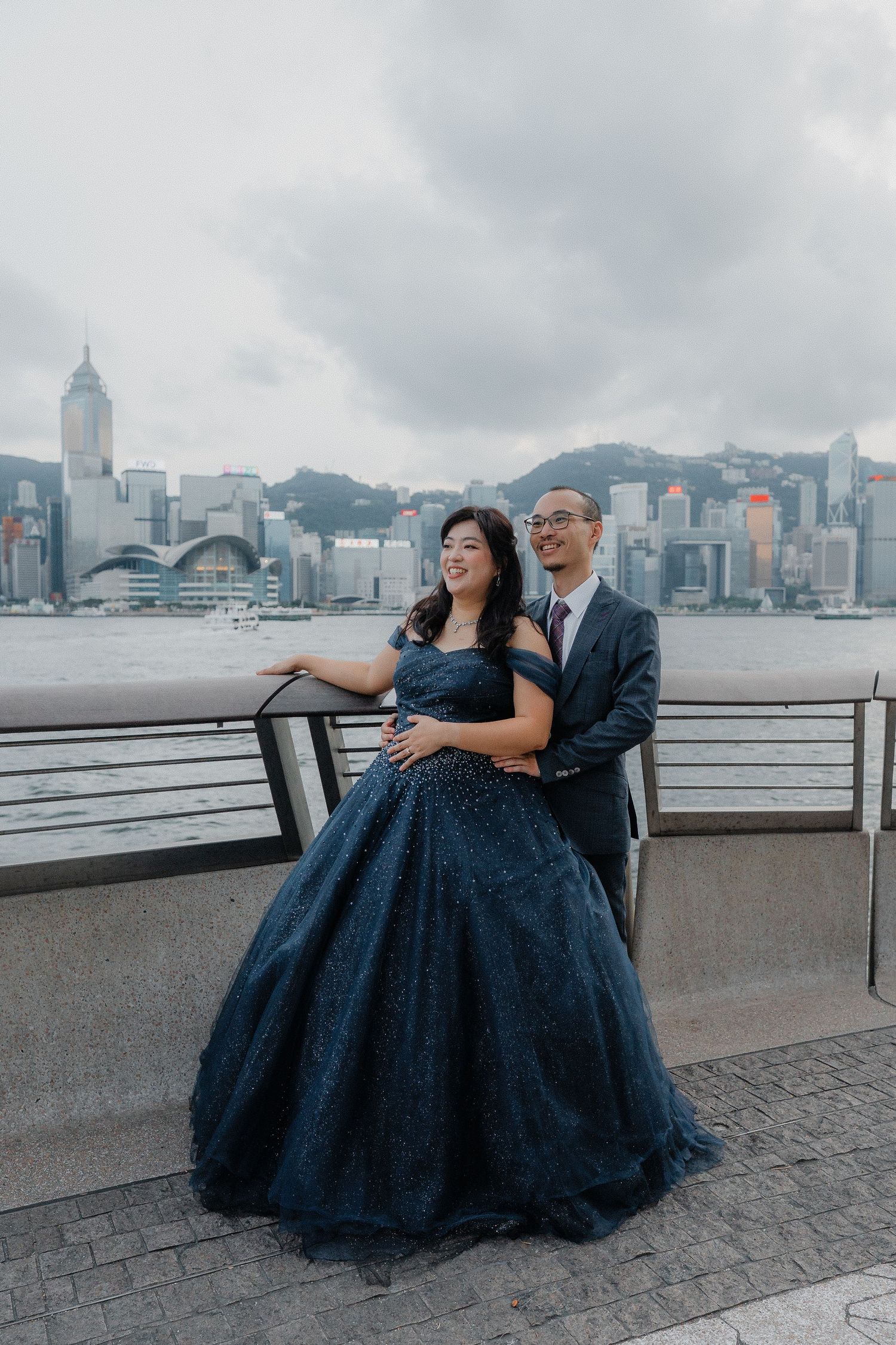 A romantic photoshoot against Hong Kong's Victoria Harbour features a couple sitting on a promenade under cloudy skies.