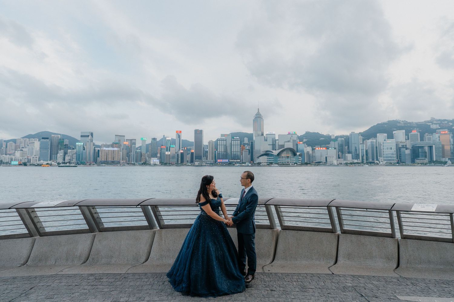 A couple in formal attire stands along Victoria Harbor waterfront with Hong Kong's iconic skyline in the background.