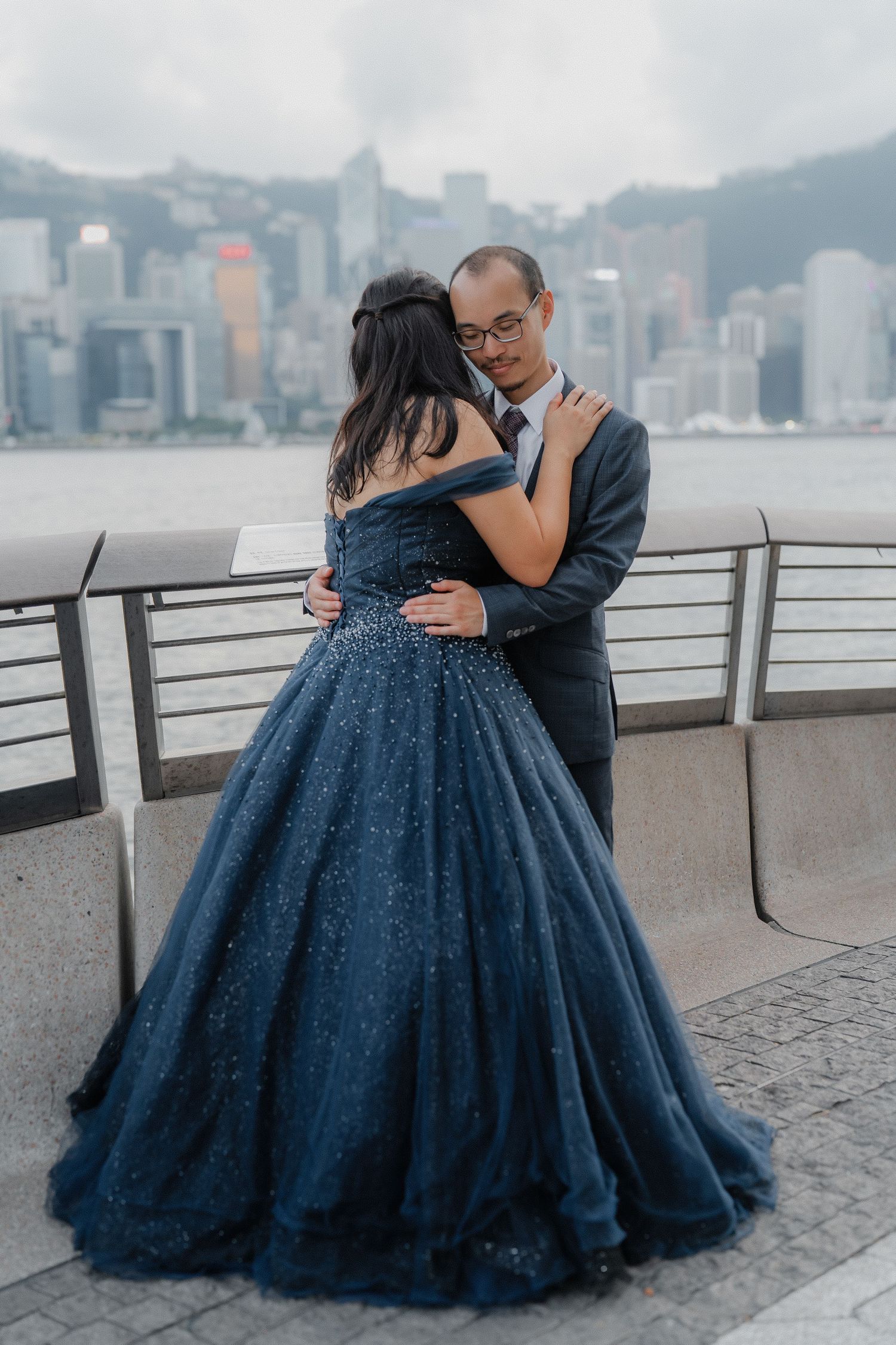A couple embraces near the Hong Kong harbor with city skyline in the background while wearing formal attire.