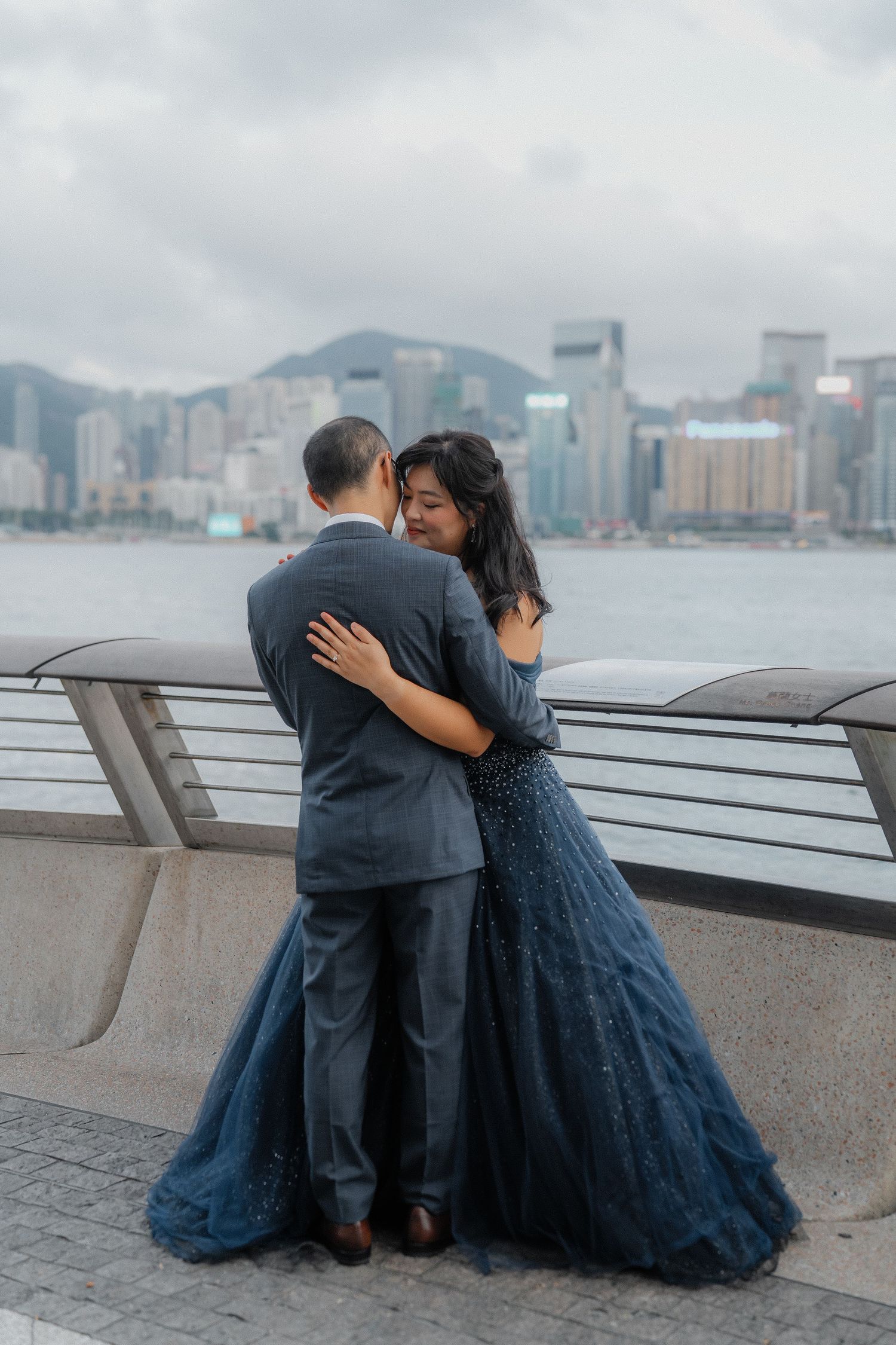 A romantic couple embraces while sitting along Hong Kong's harbor with the cityscape in the background.