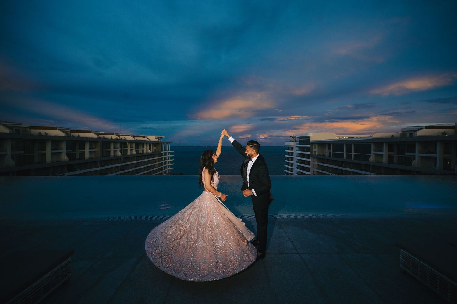 A couple in formal wedding attire dances on a deck at sunset with dark moody skies and orange clouds behind them.