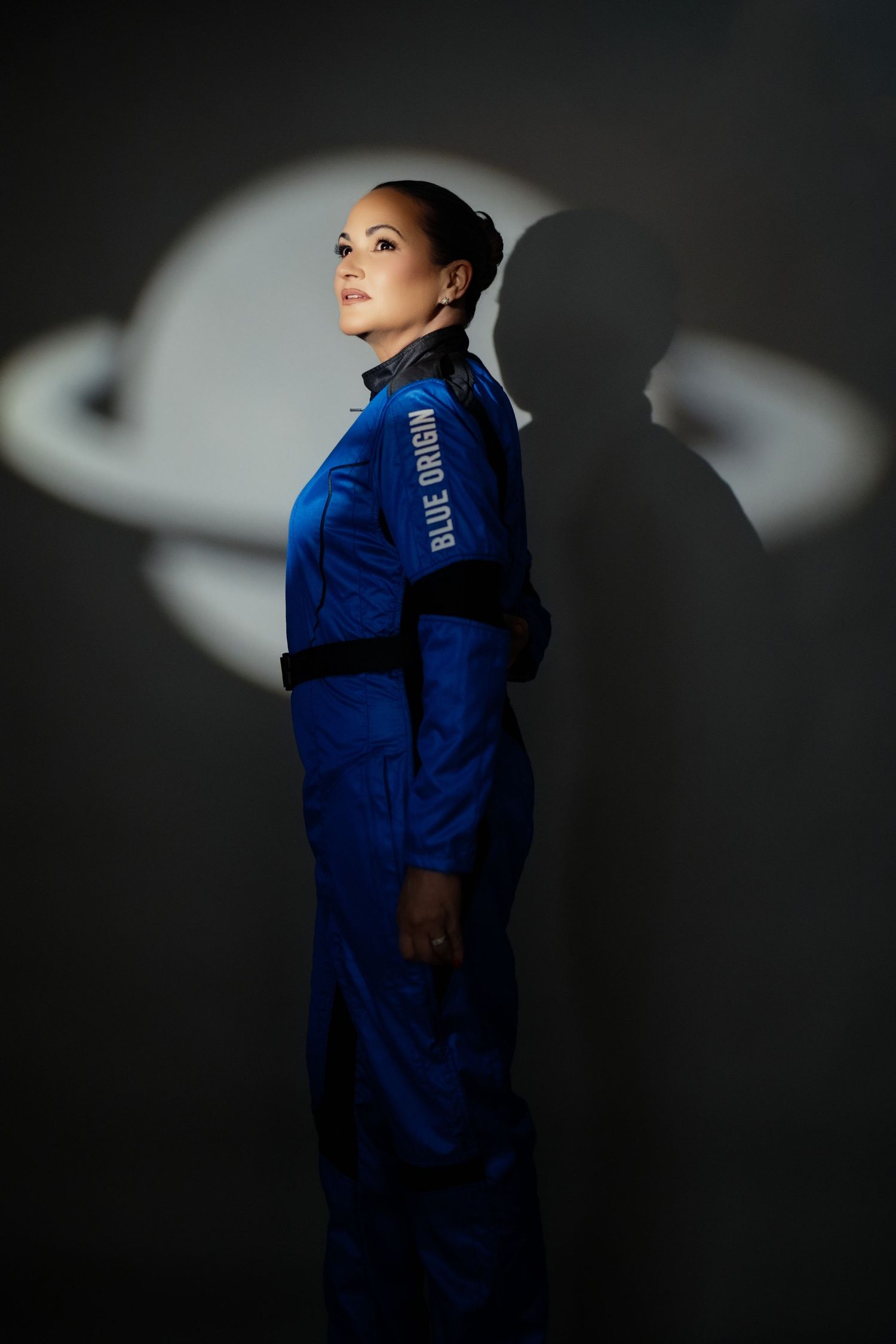 An astronaut in a blue flight suit stands in dramatic lighting with a planet-like shadow projected on the dark wall behind.