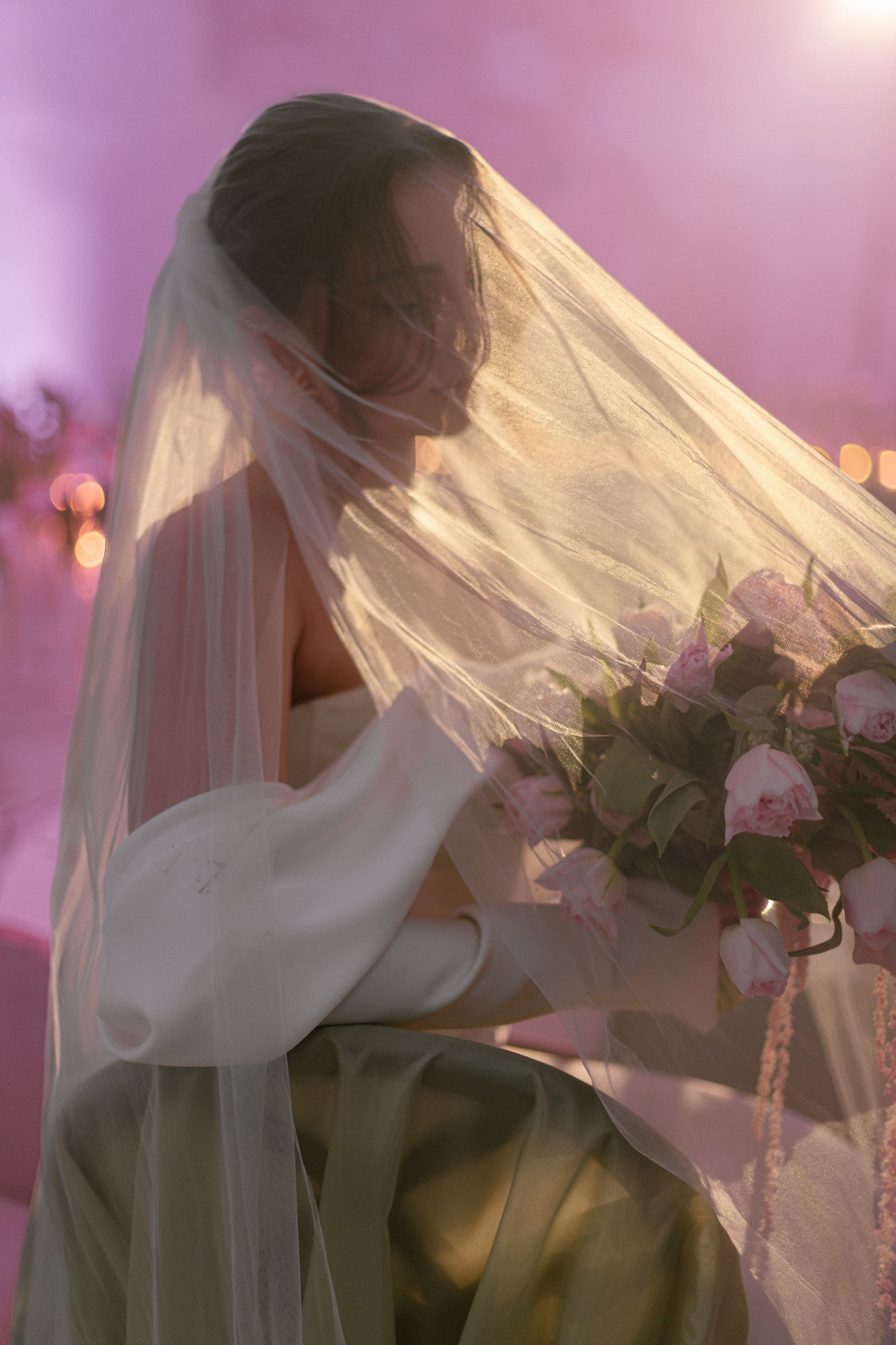 A bride in a flowing white veil holds a bouquet of pink roses against a dreamy pink-lit background.