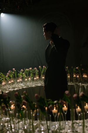 A silhouette in dark attire stands dramatically beside elegant glass vases with flowers and glowing candles.