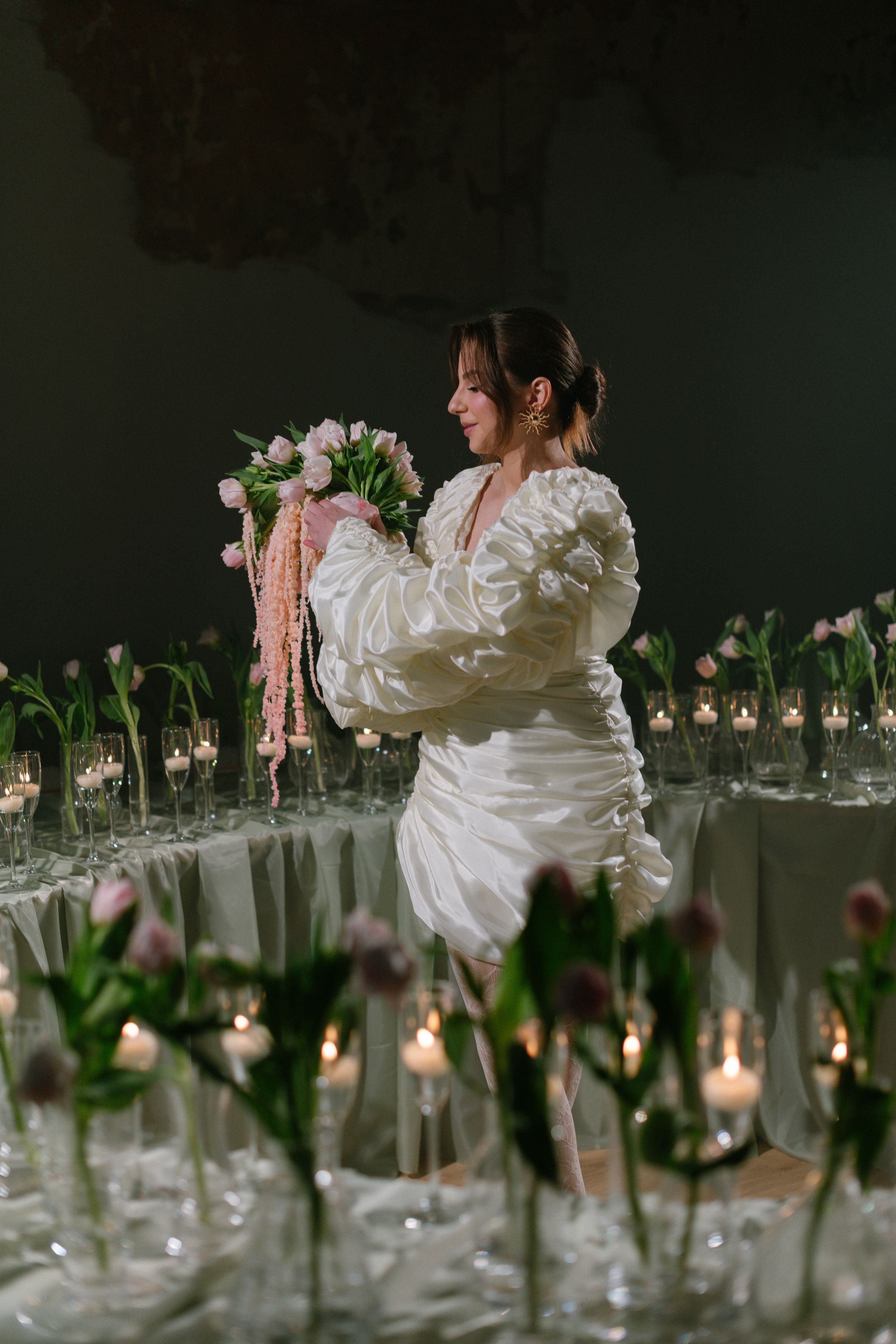 A person in an elegant white dress holds a bouquet amid a romantic table setting with candlelit floral arrangements.