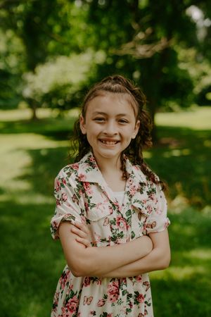 Child in floral dress stands with crossed arms and cheerful expression in park.