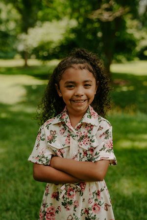 Child in floral dress stands confidently with crossed arms in a sunny garden.