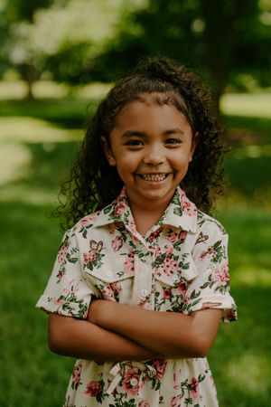 Young person in floral print dress poses with crossed arms and bright smile outdoors.
