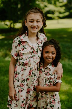 Children in coordinated floral dresses embrace while laughing in a sunny park.