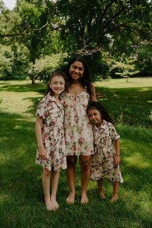 Three people wearing matching floral dresses pose together on a sunny grassy hill in a park.