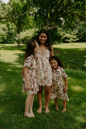 Three people in coordinating white and pink floral dresses smile together in a lush green park setting.
