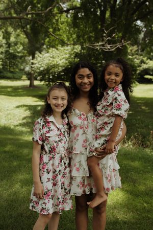 Three children in matching floral dresses smile together in sunlit park.