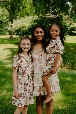 Three people wearing coordinated floral dresses laugh together on a grassy hill.