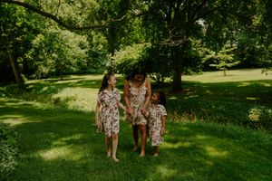 Three people in matching floral dresses stand together on green grass under trees.