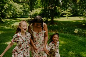 Three people in matching floral dresses share laughs together in a sunny park setting.