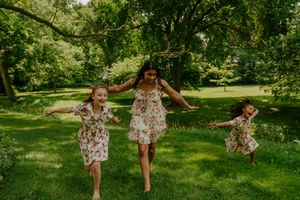 Three people in coordinated floral dresses dance and jump playfully on green grass.