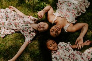 Three people in matching floral dresses lay on grass forming a circle with their heads together.