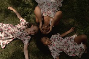 Overhead view captures three people in floral dresses making playful faces while lying in grass.