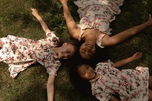 Two children in matching floral dresses play near a large branching tree in a grassy field.