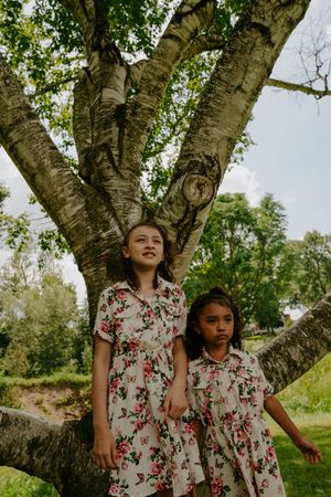 Two children wearing matching floral dresses stand beneath a large branching tree.