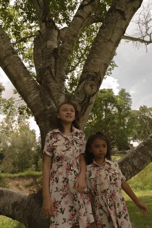 Two children in matching floral dresses explore a large branching tree in a summer meadow.