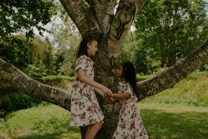 Children in matching floral dresses play around a large tree trunk in dappled sunlight.