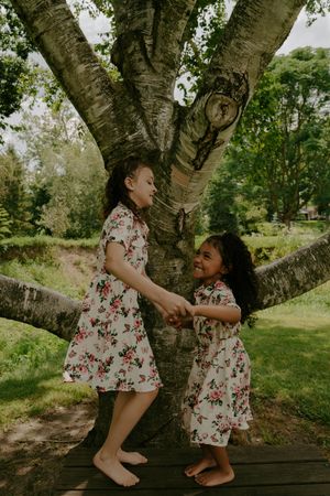 Two children in floral dresses share a playful moment on a wooden platform beneath a large tree.