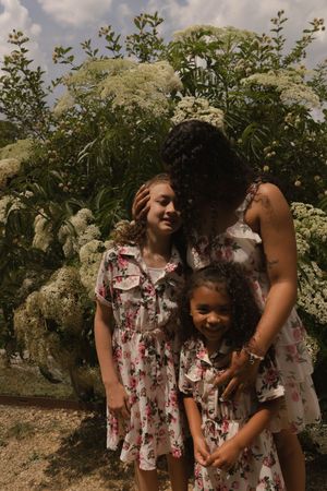 People in floral outfits stand near blooming elderflower bushes on a summer day.