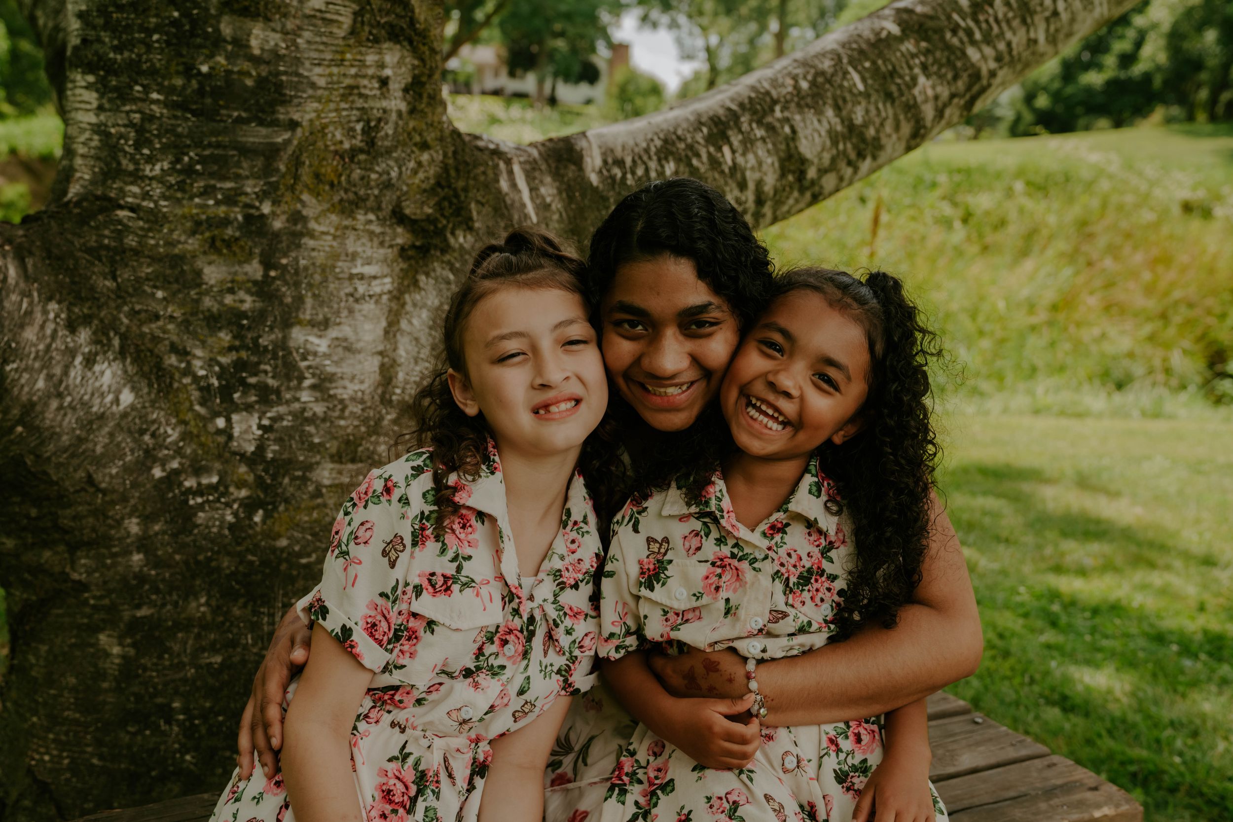 Two children in matching floral outfits share a joyful embrace against a tree trunk background.