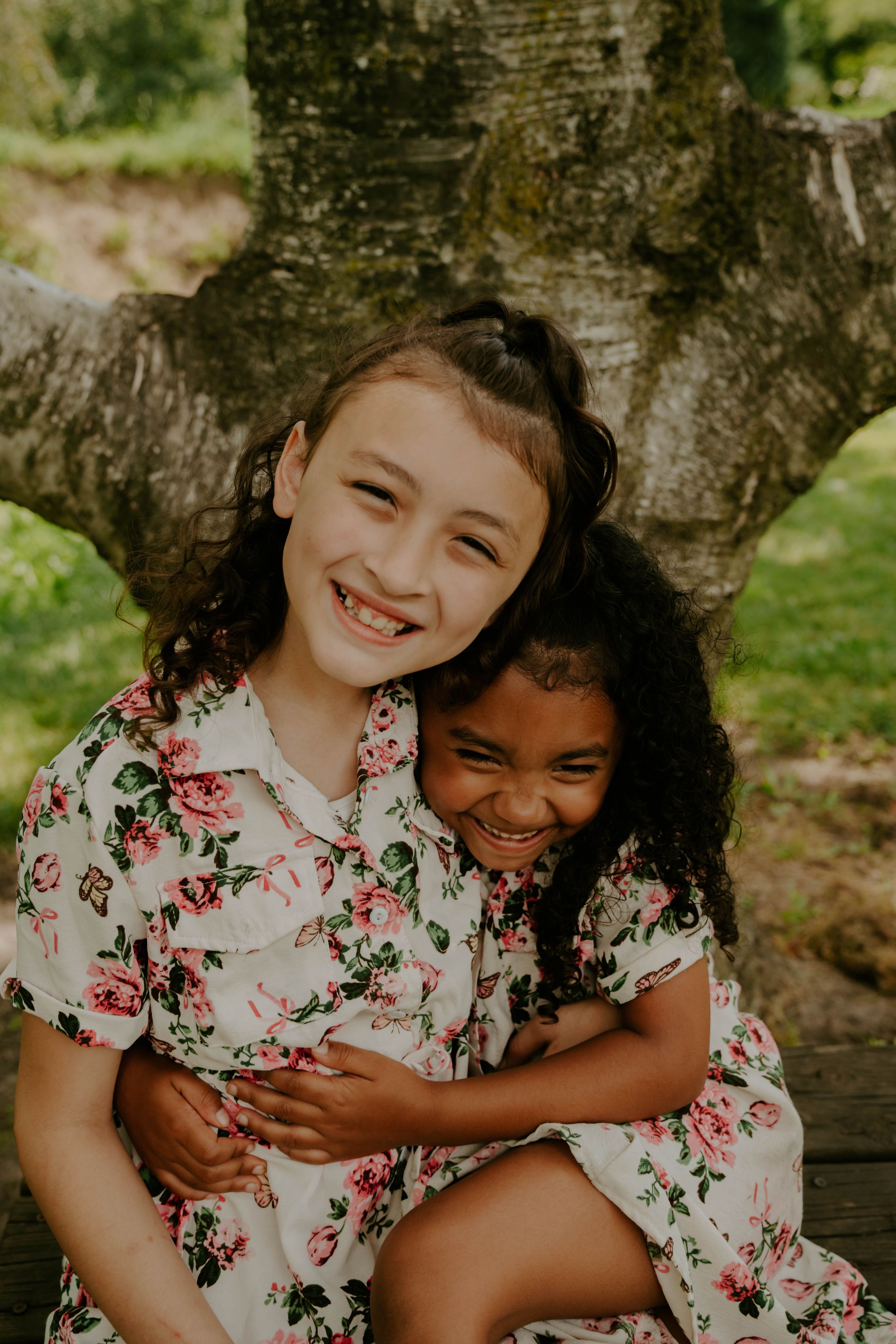 Children wearing matching floral shirts share playful moments by a large tree trunk.